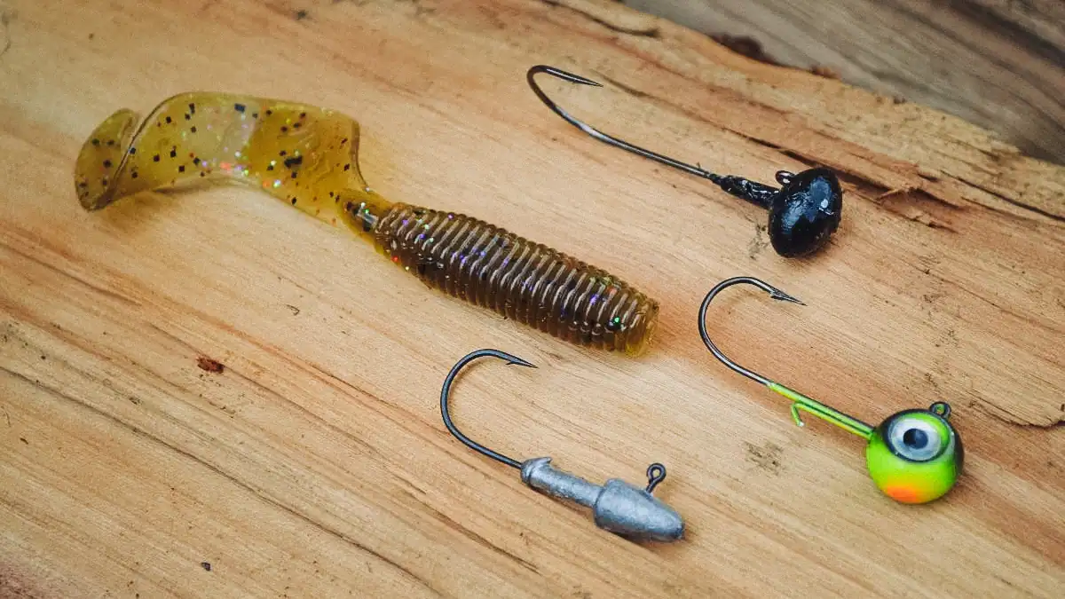 a curly tail grub on a wood table with hooks and jig heads
