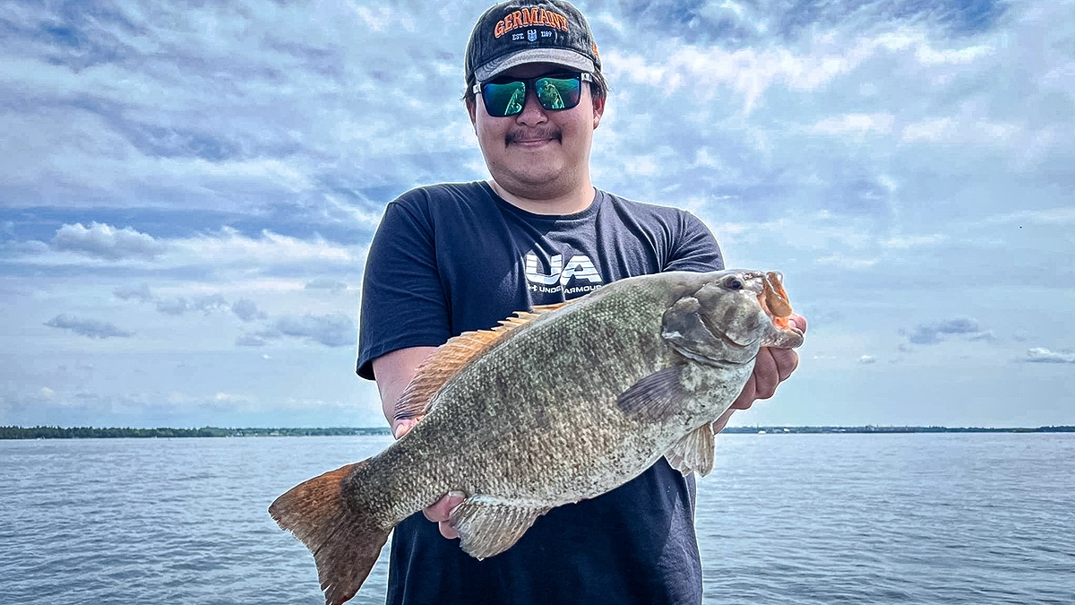 A man holds a largemouth bass with the blue water and sky behind them.