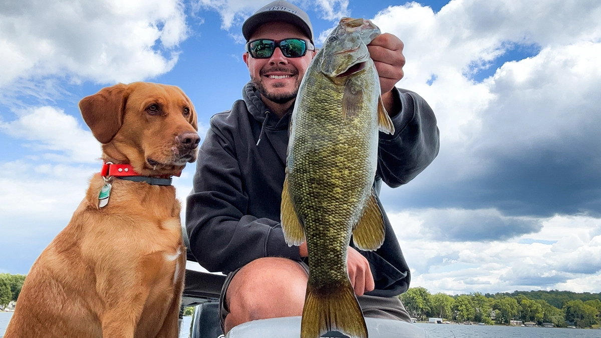 A man on the water holds a fish with a dog sitting beside him.
