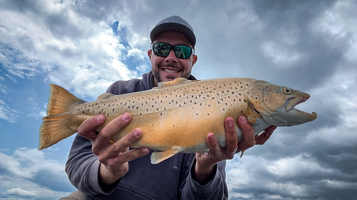 A man holds a trout with a cloudy sky dominating the background.
