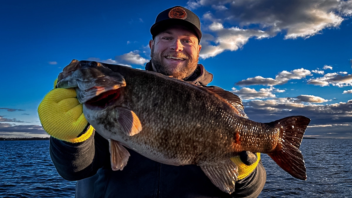 A wintertime fisher holds a bass, with the blue sky and water behind them.