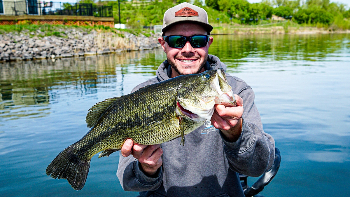 A man holds a bass near the rocky and tree-lined shore behind them