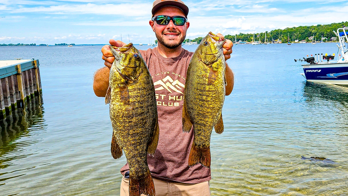 A man holds up two bass with a dock to the left, a boat to the right, and the water extending far to the background