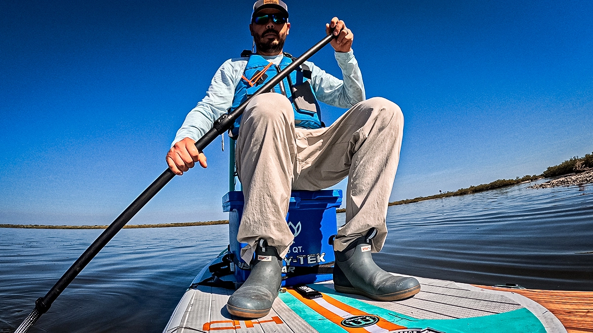 A man sits on a BOTE HD Aero SUP, with a paddle in the water and the water, shoreline and sky behind him.