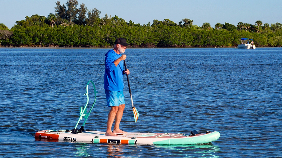 A man stands on a BOTE HD Aero SUP while holding a paddle, with trees off in the distance
