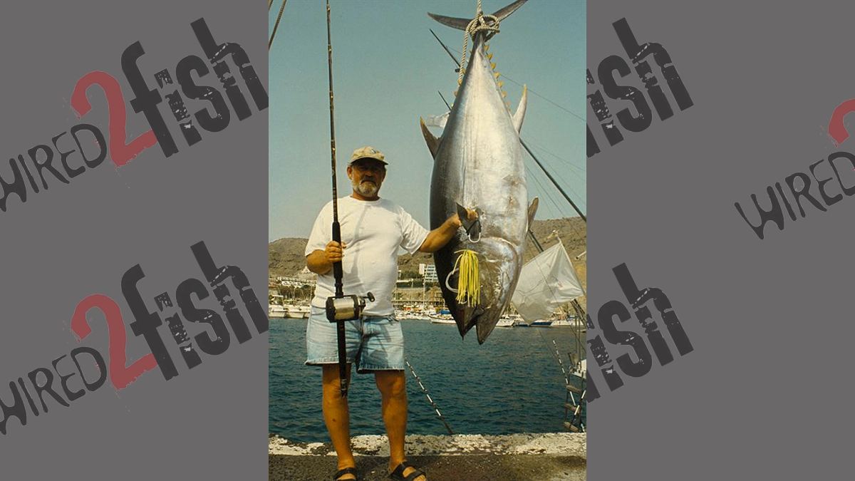 A man stands next to his world-record Atlantic bigeye tuna, with the water and shoreline behind them both.