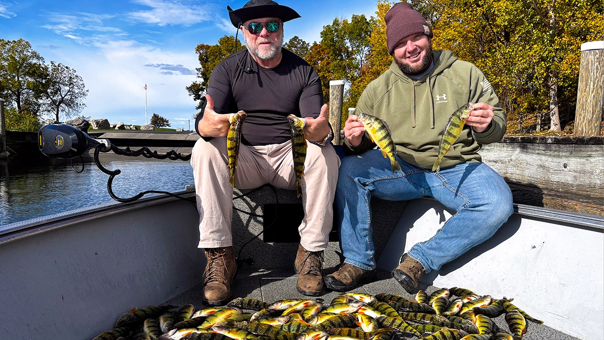 Two men on a docked boat each hold up two fish, with a pile of fish at their feet