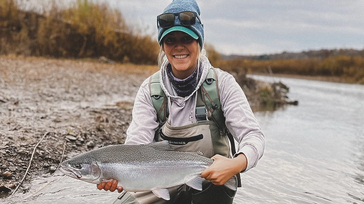 A woman holds a trout with water and a late-fall landscape in the background.