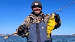 A man on the water holds up a bright yellow perch, with the shoreline far in the background.