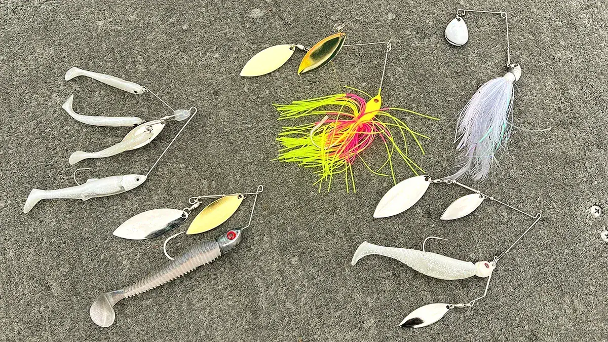 Five spinnerbaits laid out on a deck: the McCoy V-Twin Spinnerbait; a 6th Sense Flock Blade Single Colorado Spinnerbait; a Flatlands P.T. Flash Spin Spinnerbaits Triple Willow; the LIVETARGET Baitball Spinner Rig; and a Jewel Baits Gem Blade Spinnerbait.