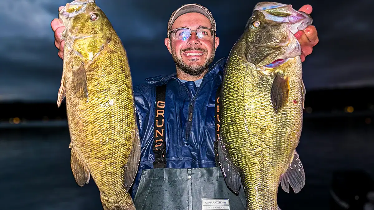 A man holds up two bass at night.