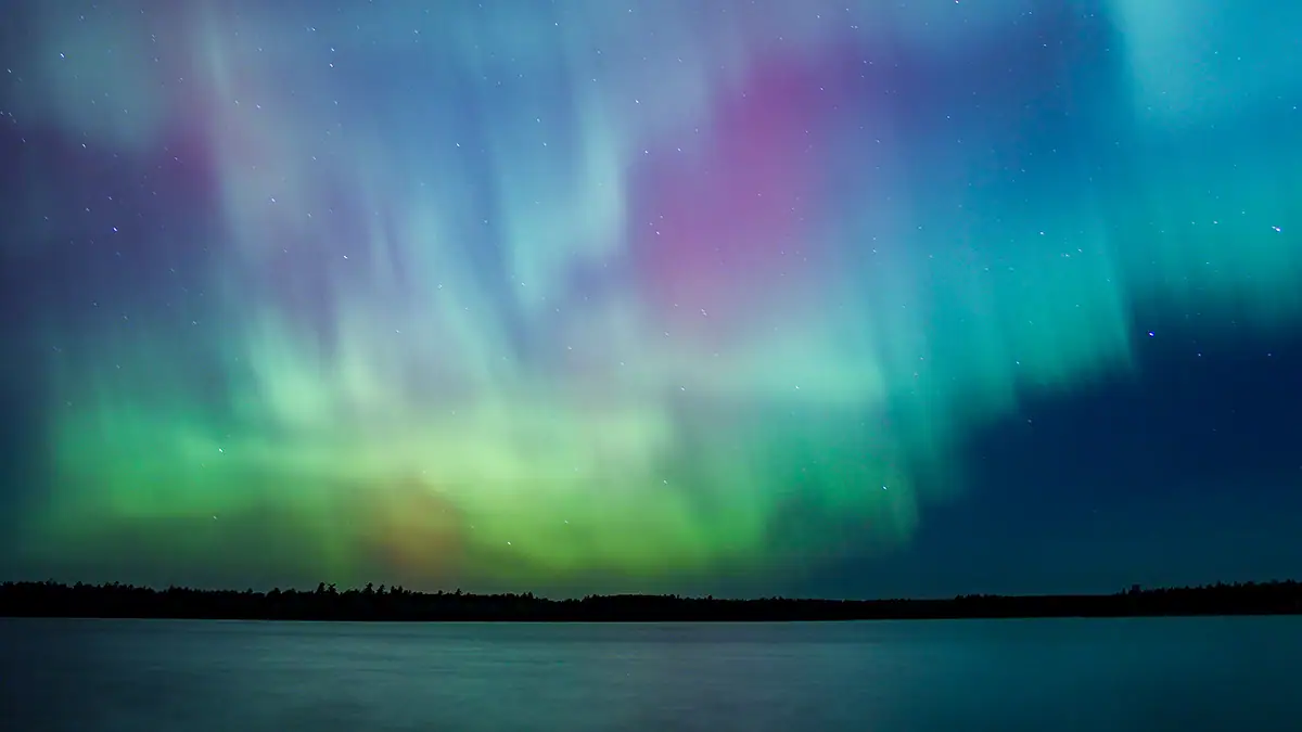 Northern lights erupt over remote Minnesota lake at night with rainbow of color shining off water Aurora Borealis nature show.